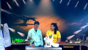 Een foto presentator Jurre en preparateur Yasmin Grooter. Op de tafel voor hen ligt de schedel van een dinosauriër en staat een opgezette Darwinvink. Op de achtergrond hangt een scherm met een animatie van een grote dinosaurus te zien, de T-Rex. 
