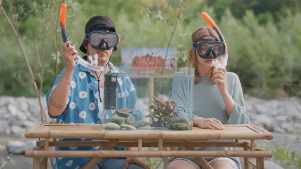 Een foto van presentatoren Jurre en Emma die met een duikbril en snorkel op achter een aquarium zitten die op een tafel staat.