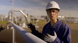 Een foto van presentator Winfried die voor de camera naast een gasleiding staat van een gas bedrijf. Hij heeft een helm en veiligheidsbril op en heeft een overall aan.