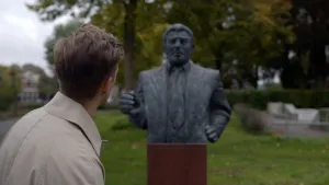 Een foto van presentator Winfried die buiten staat met zijn rug naar de camera toe. Hij kijkt naar een standbeeld van een man. Achter het standbeeld is een park te zien waar veel bomen in staan.