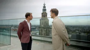 Een foto van presentator Winfried die met een man op een dak staan van een gebouw. Op de achtergrond is de Martinitoren zichtbaar, dat is een hoge stenen toren.