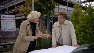 Een foto van presentator Winfried die samen met een vrouw buiten een plattegrond bespreekt. Ze staan met de rug toe naar een bouwplaats waarvan ook het hek te zien is.