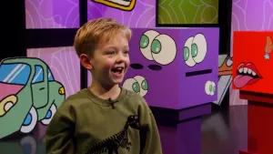 Een foto van een lachende kandidaat in de studio van het 'Raad eens wat ik doe' spel. Achter hem zijn de kijk en proef dozen te zien.