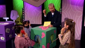 Een foto van presentator Daan die in de studio staat van het 'Raad eens wat ik doe' spel en samen met drie kandidaten rondom de hoor doos staan. Elke kandidaat luistert met een telefoon hoorn naast hun oor.