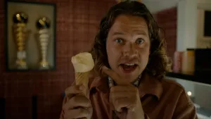 Een foto van presentator Pascal die in een ijswinkel staat . Hij verteld wat in de camera terwijl hij een ijshoorntje vasthoudt en met de wijsvinger van zijn andere hand ernaar wijst.