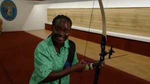 Een foto van presentator Tirsa die in een ruimte staat en lacht naar de camera. Met haar linkerhand houdt zij een ongespannen handboog vast en met haar rechterhand houdt zij een pijl vast die al op de boog klaar zit om mee te schieten. 