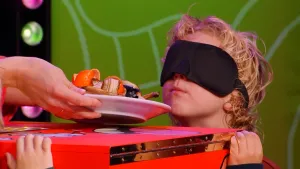 Een foto van een kandidaat in de studio van het 'Raad eens wat ik doe' spel die een blinddoek op heeft. Voor zijn gezicht wordt een bord vol met oude fruitschillen gehouden die hij moet ruiken.