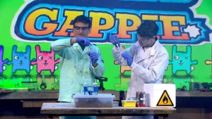 Een foto van presentator Jurre en bioloog Matyas Bittenbinder in laboratoriumkleding die aan een tafel een experiment uitvoeren. Jurre heeft een buisje met zijn eigen bloed vast, Matyas haalt slangengif uit een ander buisje met een pipet.