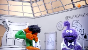 Een foto van een oranje pop die voorover gebogen in een kapotte pot zit met de handen voor het gezicht in het museum voor gewone dingen. Rechts staat er een paarse pop die naar de camera kijkt.