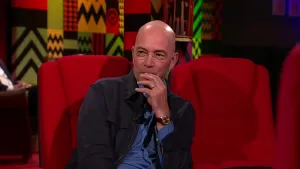 Een foto van een man die op een rode stoel zit in de studio. Hij houdt de wijsvinger van z'n linkerhand tegen zijn mond.