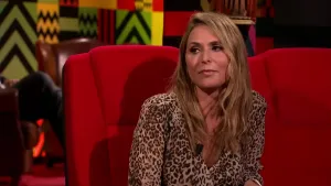 Een foto van een vrouw die op een rode stoel zit in de studio. De rode stoel naast haar is leeg en achter haar zijn de benen te zien van iemand die toekijkt.