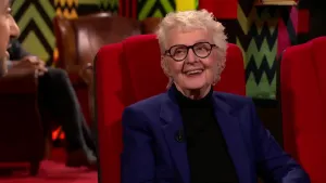 Een foto van een vrouw die op een rode stoel in de studio zit. Links is een deel van het gezicht van de presentator te zien.