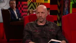 Een foto van een man die op een rode stoel zit in de studio. Achter hem zit er een man die toekijkt.