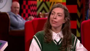 Een foto van een vrouw die op een rode stoel zit in de studio. Achter haar zit een man die toekijkt.