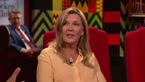 Een foto van een vrouw die op een rode stoel zit in de studio. Achter haar kijkt een man toe.
