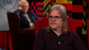 Een foto van een man die een bril draagt die op een rode stoel zit in de studio. Achter hem kijkt er een vrouw toe terwijl ze leunt op haar elleboog.
