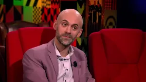 Een foto van een man die op een rode stoel zit in de studio. De rode stoel naast hem is leeg.