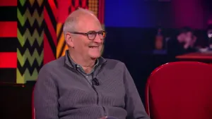 Een foto van een man die lachend in een rode stoel zit in de studio. De rode stoel naast hem is leeg.
