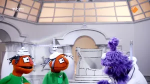 Een foto van twee oranje poppen en een paarse pop in een zwart-witte kamer. De paarse pop praat tegen de oranje poppen. Alle dingen in de kamer hebben zwarte lijntjes alsof ze getekend zijn. 