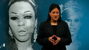 Een foto van een presentator met op de achtergrond een foto van twee leden van de LHBTQ-gemeenschap in New York.