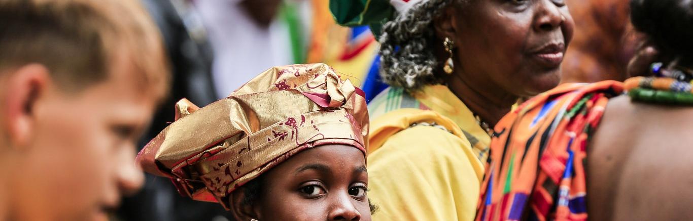 Kind met traditioneel Surinaams hoofddeksel tussen andere Surinaams geklede mensen tijdens optocht Keti Koti