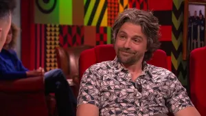 Een foto van een man die op een rode stoel in de studio zit. Links is er een deel van het gezicht van de presentator te zien. Op de achtergrond zit er een vrouw die toekijkt.