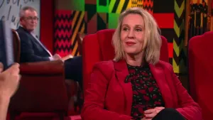 Een foto van een vrouw die op een rode stoel in de studio zit. Links is er een hand te zien die een boek vasthoudt en op de achtergrond zit er een man die toekijkt. 