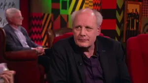 Een foto van een man die op een rode stoel in de studio zit. Op de achtergrond is er nog een man te zien die in een stoel zit.