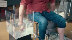 Een foto van presentator Diederik Jekel die met z'n handen en voeten elk in bakken met water zit. Met zijn handen houdt hij een metaal plaatjes vast waar draden aan vastzitten.