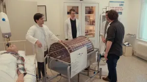 Een foto van presentator Diederik die met drie mannen rondom een kunstnier staan. Het kunstnier is een cilinder van twee fietswielen die aan elkaar vastzit met een rol van metalen delen. Er komt een slang uit die naar een pop gaat die op bed ligt.