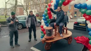 Een foto van presentator Diederik Jekel die bovenop een houten elektrische auto zit die onder een boog van ballonnen door rijdt.
