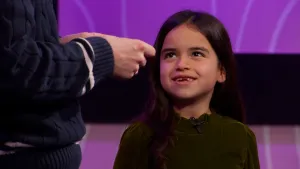 Een foto van een kandidaat van het 'Raad eens wat ik doe' spel. Zij staat tegenover presentator Daan waarvan alleen zijn armen te zien zijn.