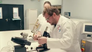Een foto van presentator Diederik Jekel die in een laboratorium bezig is met een microscoop. Naast hem staat er een vrouw en er staat nog een microscoop met verschillende apparaten op tafel.