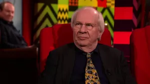 Een foto van een man die op een rode stoel in de studio zit. Achter hem zit een man die toekijkt.