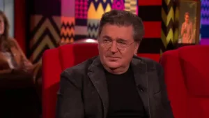 Een foto van een man die op een rode stoel in de studio zit. Achter hem zit een vrouw die toekijkt.