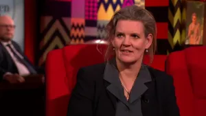 Een foto van een vrouw die op een rode stoel in de studio zit. Op de achtergrond zit er een man die toekijkt.