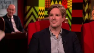 Een foto van een man die in een studio zit op een rode stoel. Op de achtergrond zit er nog een man die toekijkt.