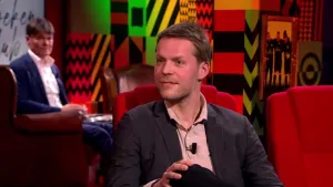 Een foto van een man die in de studio zit op een rode stoel. Op de achtergrond zit er nog een man toe te kijken.