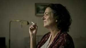 Een foto van een vrouw die in een huiskamer een sigaret aan het roken is. Op de achtergrond is er een lamp, bank en een schilderij zichtbaar.