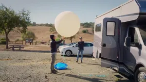 Een foto van een man die buiten een grote witte ballon vult met gas. De slang van de ballon loopt een camper in. Op de achtergrond staat er een man toe te kijken.
