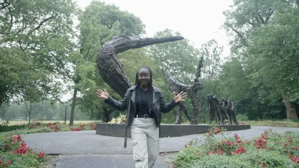 Een foto van presentator Tirsa die wat uitlegt voor de camera over het slavernijmonument op de achtergrond.