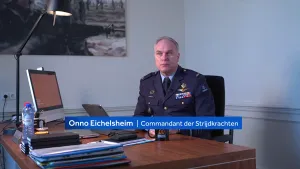 Een foto van een commandant die in zijn kantoor zit achter een bruin bureau. Voor hem staan twee computers en een gesloten laptop.