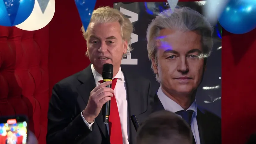 Een foto van Geert Wilders die praat met een microfoon in zijn hand. Boven hem hangen er blauw en witte slingers en ballonnen. Op de achtergrond is er een grote foto van zijn gezicht en de naam van zijn politieke partij.