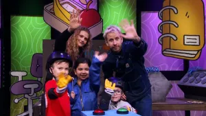 Een foto van presentator Daan die aan het einde van het 'Raad eens wat ik doe' spel samen met drie kandidaten en een vrouw die motoren repareert gedag zwaaien in de camera.