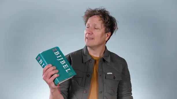 Een foto van een presentator die een bijbel in zijn rechterhant heeft en naar de bijbel kijkt. Op de bijbel is de opdruk "De Bijbel" leesbaar.