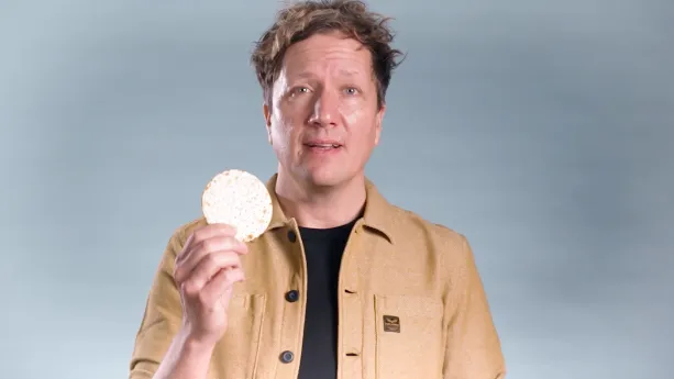 Een foto van een presentator die in de camera kijkt en een kleine Matze omhoog houdt. Een Matze is een soort ronde cracker.