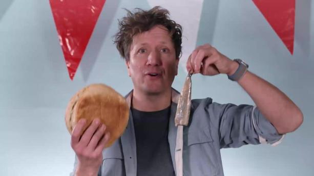 Een foto van presentator Klaas met in zijn handen haring en wit brood. Boven hem hangen vlaggen.