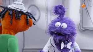 Een foto van een oranje pop en een paarse pop. De oranje pop staat met de rug naar de camera en kijkt naar de paarse pop. Ze staan in een zwart-witte kamer.