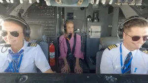 Een foto van twee piloten in de cockpit van een vliegtuig. Ze dragen allebei een headset. Om hen heen zijn heel veel knopjes te zien. Achter hen zit Presentator Janouk in het gangpad.