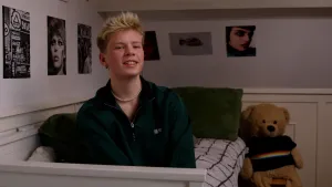 Een foto van Pim die op zijn bed zit. Op de achtergrond zijn kleine posters zichtbaar van mensen, en een straat vol borden. Naast het bed staat een grote knuffel van een beer die een zwarte trui met een kleurige lijn erop aanheeft.